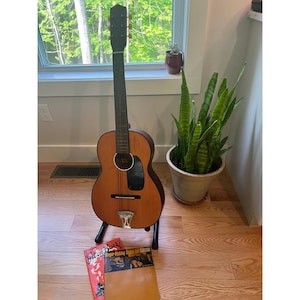 Puede incluir: Guitarra acústica con acabado de madera natural, expuesta sobre un soporte negro. La guitarra tiene mástil y clavijero oscuros, con cordal plateado. Un álbum vintage titulado "Get on Board" es visible en primer plano. Una planta de serpiente en maceta está en el fondo.