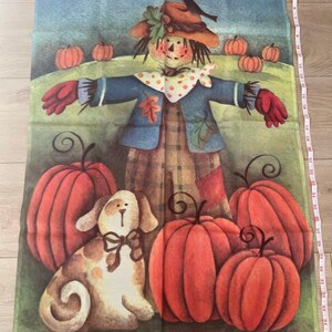 May include: A decorative fall-themed kitchen towel featuring a scarecrow with pumpkins and a dog. The scarecrow wears a blue jacket, plaid overalls, and a hat. The pumpkins are orange, and the dog is tan with brown spots. The towel measures approximately 23 inches in length.