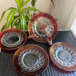 May include: A set of vintage amber glass plates and a serving dish. The plates have a clear center surrounded by a ring of small, raised circles. The serving dish has wooden handles. The glass has a reddish-brown hue.