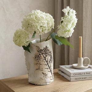Op de afbeelding: Een crèmekleurige keramische vaas met een boomontwerp bevat witte hortensia's. Een kleine witte kandelaar met een brandende kaars en twee boeken staan op een houten oppervlak. De vaas heeft een ruwe, getextureerde rand.