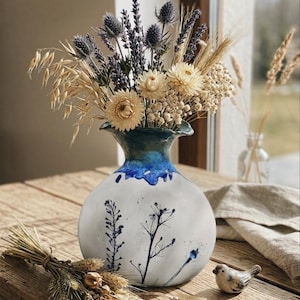 Może przedstawiać: Biały ceramiczny wazon z niebieską obwódką i kwiatowym wzorem zawiera bukiet suszonych kwiatów, w tym lawendę i pszenicę. Mały ceramiczny ptak siedzi na drewnianym stole obok wazonu. Wazon ma około 25 cm wysokości.