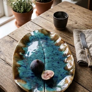 May include: A ceramic leaf-shaped plate with a teal and brown glaze, holding a whole fig and a halved fig. The plate sits on a wooden table, alongside a small dark cup, a linen napkin, and a knife. Potted plants are in the background.