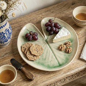 Puede incluir: Una fuente para servir en forma de hoja con un esmalte craquelado verde y crema, que contiene uvas, galletas, queso brie y nueces. También hay un cuenco pequeño de miel y un cuchillo para queso. La fuente está sobre una mesa de madera.
