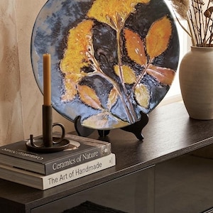 Può includere: Piatto decorativo in ceramica con un design botanico di foglie gialle e arancioni su sfondo blu scuro. Il piatto è esposto su un supporto nero, con libri e una candela nelle vicinanze.