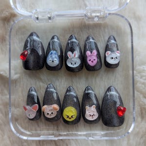 May include: A set of press-on nails in a clear plastic case. The nails are dark gray with a glittery finish and feature various cartoon animal designs and red heart-shaped gems. The nail shapes are almond-shaped.