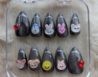 May include: A set of press-on nails in a clear plastic case. The nails are dark gray with a glittery finish and feature various cartoon animal designs and red heart-shaped gems. The nail shapes are almond-shaped.