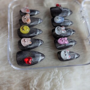 May include: A clear plastic case holds a set of press-on nails. Each nail is a dark gray color with a marbled effect and adorned with a small, colorful cartoon animal or gem. The animals include a pink pig, a white bunny, and a yellow chick.