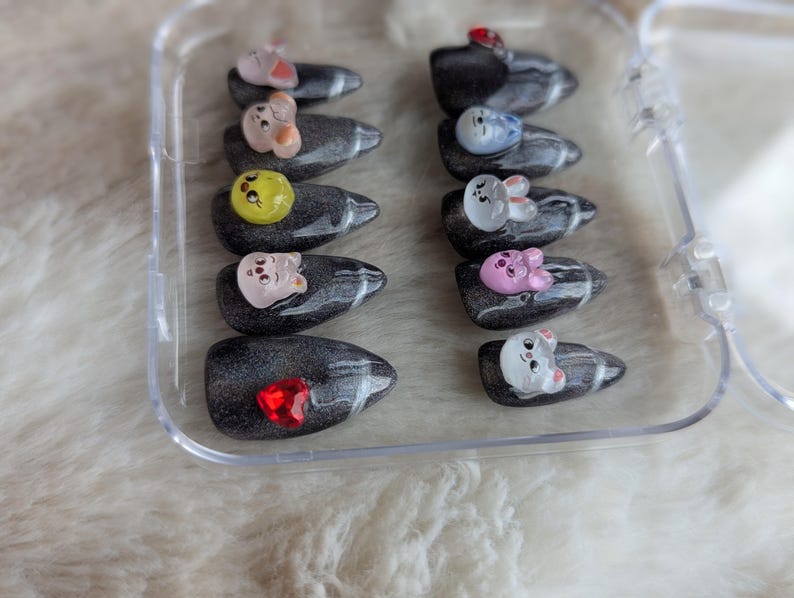 May include: A clear plastic case holds a set of press-on nails. Each nail is a dark gray color with a marbled effect and adorned with a small, colorful cartoon animal or gem. The animals include a pink pig, a white bunny, and a yellow chick.