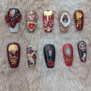 Pode incluir: Dez unhas postiças em forma de caixão vermelhas e douradas com vários desenhos temáticos de Alice no País das Maravilhas, incluindo uma xícara de chá, um relógio, um cartão de jogo e um coração.