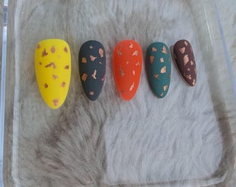May include: Five false nails with a long oval shape. Each nail is painted a different color: yellow, dark green, orange, light green, and brown. Each nail has a design of small gold flecks.