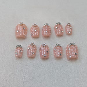 Pode incluir: Um conjunto de dez unhas artificiais em tom rosa claro, adornadas com bolinhas brancas. Cada unha apresenta um adorno decorativo na base e pequenas pedras brilhantes. As unhas estão dispostas sobre uma superfície clara.