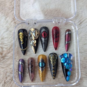 May include: A set of 10 false nails with different designs. The nails are in a clear plastic case. The designs include a snake, a dragon, an eye, a flower, and a geometric pattern. The nails are black, gold, red, and blue.
