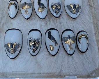 May include: A set of ten white oval press-on nails with black edges. Each nail features a different design, including gold and silver moth, bird, and crescent moon accents. The nails are arranged in a clear plastic container.