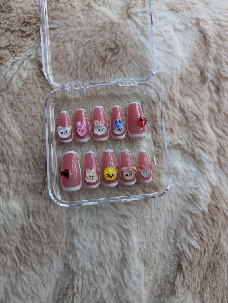 May include: A set of press-on nails in a clear plastic case. The nails are a light pink with cartoon animal designs and a red heart-shaped gem. The nails are rectangular in shape.