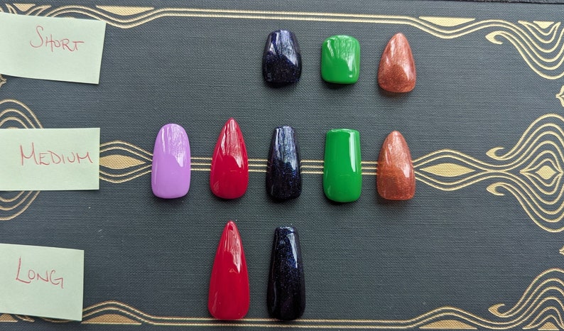 May include: A collection of press-on nails in various colors and shapes, arranged by length: short, medium, and long. Colors include purple, red, green, and a glittery dark blue and copper. The nails are displayed on a dark background with gold decorative accents.