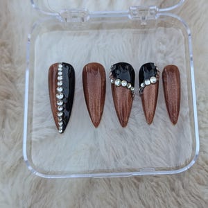 Pode incluir: Conjunto de seis unhas postiças com base de glitter marrom e pontas pretas. Cada unha tem uma fileira de strass transparentes na ponta preta.