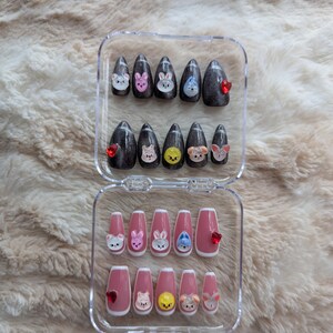 May include: A set of press-on nails in a clear plastic case. The nails feature cartoon animal faces and red heart accents. Some nails are dark gray with glitter, while others are pink with white tips.