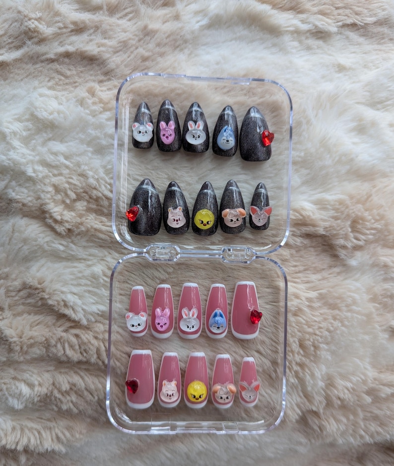 May include: A set of press-on nails in a clear plastic case. The nails feature cartoon animal faces and red heart accents. Some nails are dark gray with glitter, while others are pink with white tips.