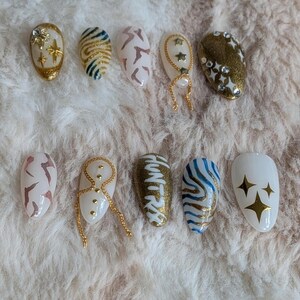 Pode incluir: Uma cole&ccedil;&atilde;o de dez unhas posti&ccedil;as ovais com v&aacute;rios designs. As unhas apresentam cores douradas, brancas e castanhas, com padr&otilde;es que incluem estrelas, riscas e formas abstratas. Algumas unhas s&atilde;o adornadas com correntes douradas e strass.