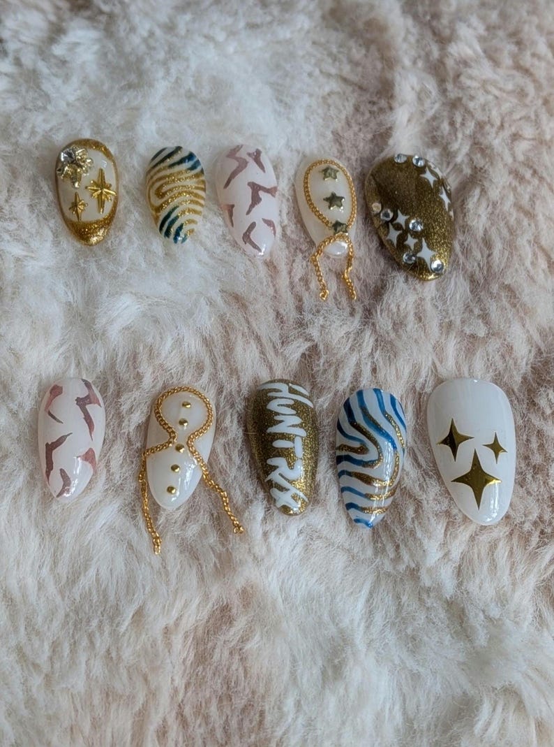 Pode incluir: Uma cole&ccedil;&atilde;o de dez unhas posti&ccedil;as ovais com v&aacute;rios designs. As unhas apresentam cores douradas, brancas e castanhas, com padr&otilde;es que incluem estrelas, riscas e formas abstratas. Algumas unhas s&atilde;o adornadas com correntes douradas e strass.