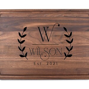 May include: A personalised walnut chopping board with a wreath design, the initial "W", the name "Wilson", and the text "Est. 2021".