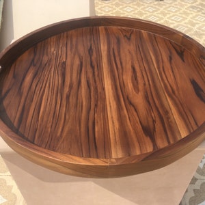May include: A large, round wooden serving tray with a natural wood finish and a raised edge. The tray has two handles on the sides.