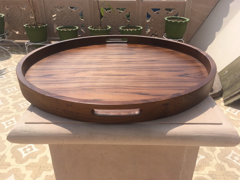 May include: A large, round wooden serving tray with two handles on the sides. The tray is made of dark brown wood with a natural wood grain pattern.