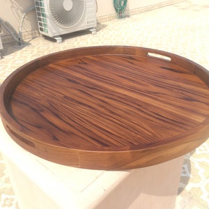 May include: A large, round wooden serving tray with two handles. The tray is made of dark brown wood with a natural grain pattern.