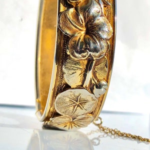 Könnte beinhalten: Ein goldenes Armreif-Armband mit floralem Design. Das Armband besteht aus zwei verschiedenen Goldtönen, wobei die Blumen einen helleren Farbton haben. Das Armband hat eine Kette daran.