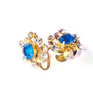 May include: A pair of gold-tone clip-on earrings featuring blue and clear rhinestones. The earrings have a floral design with a large blue rhinestone in the centre of each flower.