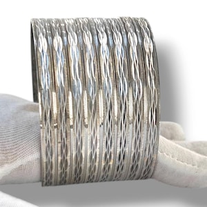 Peut inclure: Un ensemble de huit bracelets joncs en métal argenté présentant un motif gravé en forme de losanges répétitifs. Chaque bracelet possède une finition polie avec des surfaces réfléchissantes créant un aspect texturé sur l'ensemble.