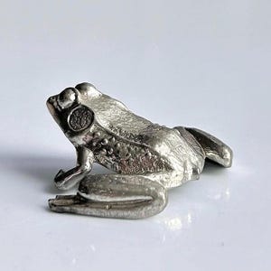 Peut inclure: Petite figurine de grenouille argentée. La grenouille est représentée en position assise, les pattes repliées. La figurine présente des textures détaillées et une finition brillante, lui donnant un aspect réaliste.
