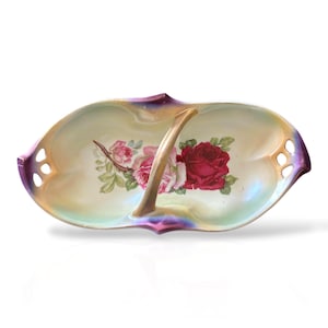 May include: Oval-shaped, divided serving dish with a central handle. The dish features a floral rose design in shades of red and pink, with green leaves. The rim has a purple and gold iridescent finish, and the dish has three small holes on each end.