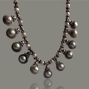 May include: A silver-toned necklace with a series of pearl pendants. The necklace features small, round pearls strung together, with larger pearl pendants hanging at intervals. The pendants are set in ornate silver settings.