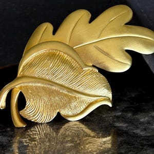 Peut inclure: Une broche dorée en forme de feuille. La broche présente deux feuilles qui se chevauchent avec des nervures détaillées et une surface texturée. Les feuilles ont une finition mate et sont posées sur un fond sombre.