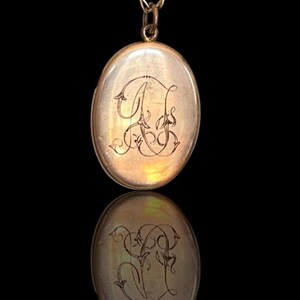 Peut inclure: Pendentif en forme de médaillon ovale en or, orné d'un monogramme gravé. Le médaillon est suspendu à une fine chaîne en or. Le monogramme présente des lettres entrelacées et ornées. La surface du médaillon reflète la lumière.