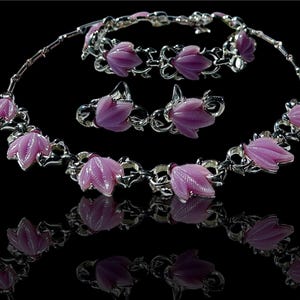 Puede incluir: Un conjunto de joyas de flores de vidrio rosa, que incluye un collar, una pulsera y pendientes. Las joyas están hechas de metal plateado y presentan delicados diseños florales.