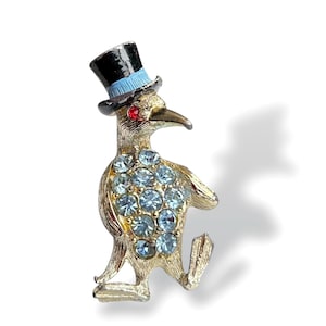 Peut inclure: Une broche en forme de pingouin dorée portant un chapeau haut de forme noir avec un ruban bleu. Le pingouin est décoré de strass bleu clair et a des yeux rouges.