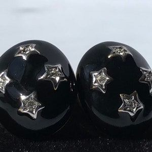 K&ouml;nnte beinhalten: Ein Paar schwarze und silberne Ohrringe mit einem Sternendesign. Jeder Ohrring hat f&uuml;nf silberne Sterne mit einem klaren Strassstein in der Mitte.