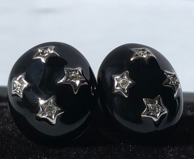 K&ouml;nnte beinhalten: Ein Paar schwarze und silberne Ohrringe mit einem Sternendesign. Jeder Ohrring hat f&uuml;nf silberne Sterne mit einem klaren Strassstein in der Mitte.