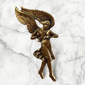 Könnte beinhalten: Eine goldfarbene Metallengelfigur mit ausgebreiteten Flügeln und einem fließenden Rock. Der Engel steht auf einer weißen Marmorfläche.