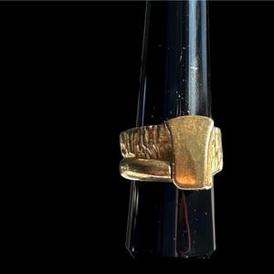 May include: A gold-colored ring with a textured band and a rectangular top. The ring is displayed on a black ring holder against a black background. The ring's design is geometric and modern.