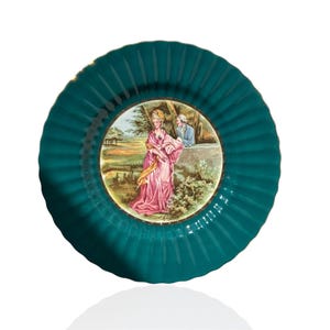 Può includere: Un piatto decorativo color verde acqua con bordo scanalato e rifiniture dorate. Il centro del piatto presenta una scena dipinta di una donna in abito rosa e un uomo in cappotto blu. Il piatto è rotondo.