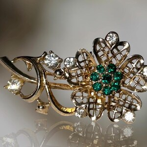 May include: A gold-tone brooch with a delicate floral design. The brooch features a four-leaf clover with a center cluster of green rhinestones and smaller clear rhinestones throughout.