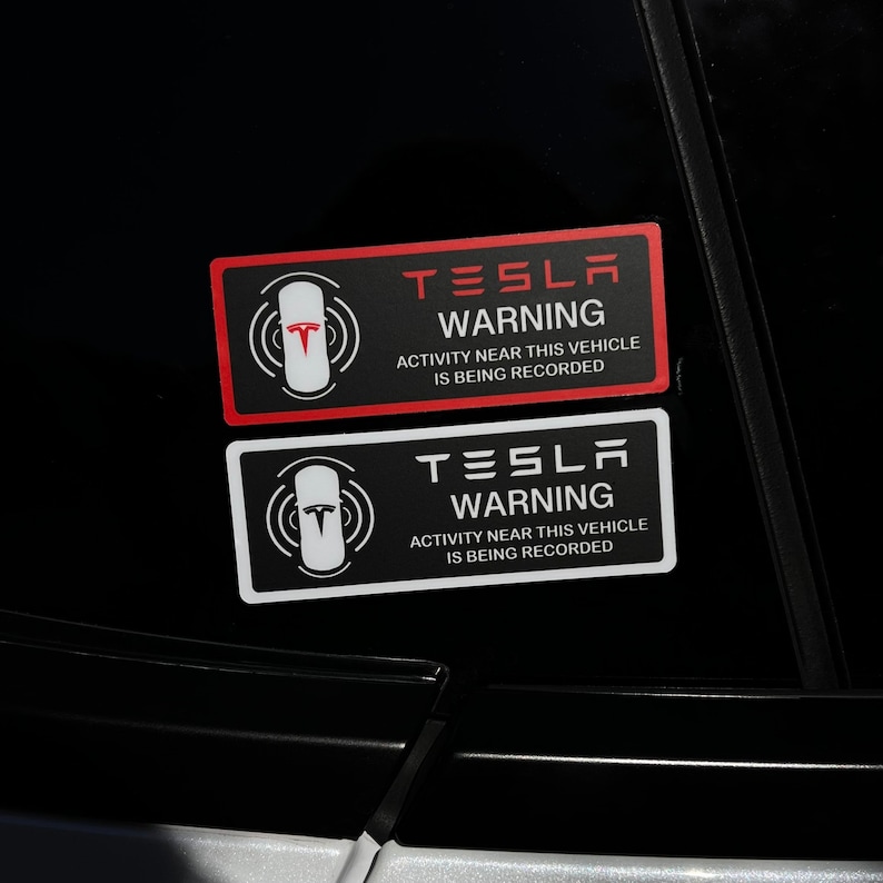 May include: Two black and white warning stickers with the text "TESLA WARNING" and "ACTIVITY NEAR THIS VEHICLE IS BEING RECORDED". The top sticker has red text and a red Tesla logo. The bottom sticker has white text and a white Tesla logo.