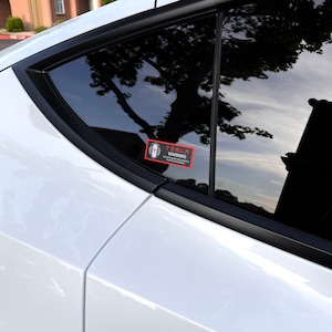 May include: A black and white warning sticker on a white car window. The sticker says "Tesla Warning" and has a symbol of a person with a hand outstretched. The text below the symbol says "Active near this vehicle.  Always use caution around vehicles."
