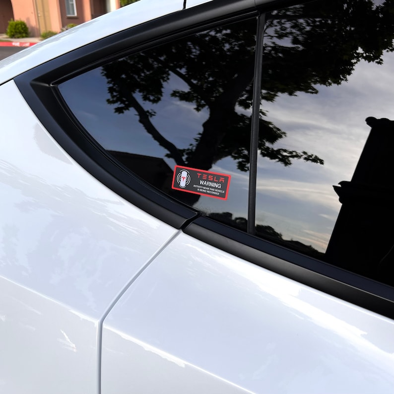 May include: A black and white warning sticker on a white car window. The sticker says "Tesla Warning" and has a symbol of a person with a hand outstretched. The text below the symbol says "Active near this vehicle.  Always use caution around vehicles."