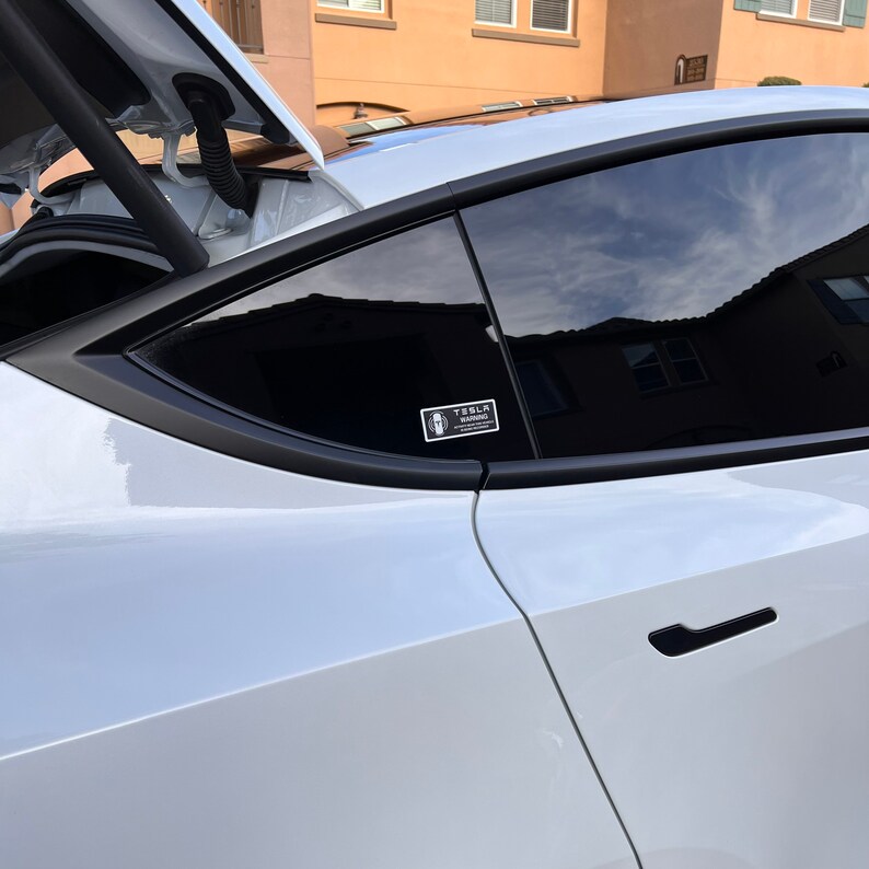 May include: A white Tesla car with a black window and a black Tesla warning sticker on the window. The car has a black door handle.