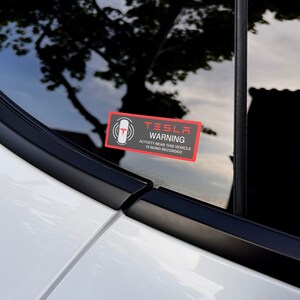 May include: A black and red warning sticker that reads "Tesla Warning" and "Activity near this vehicle is being recorded".
