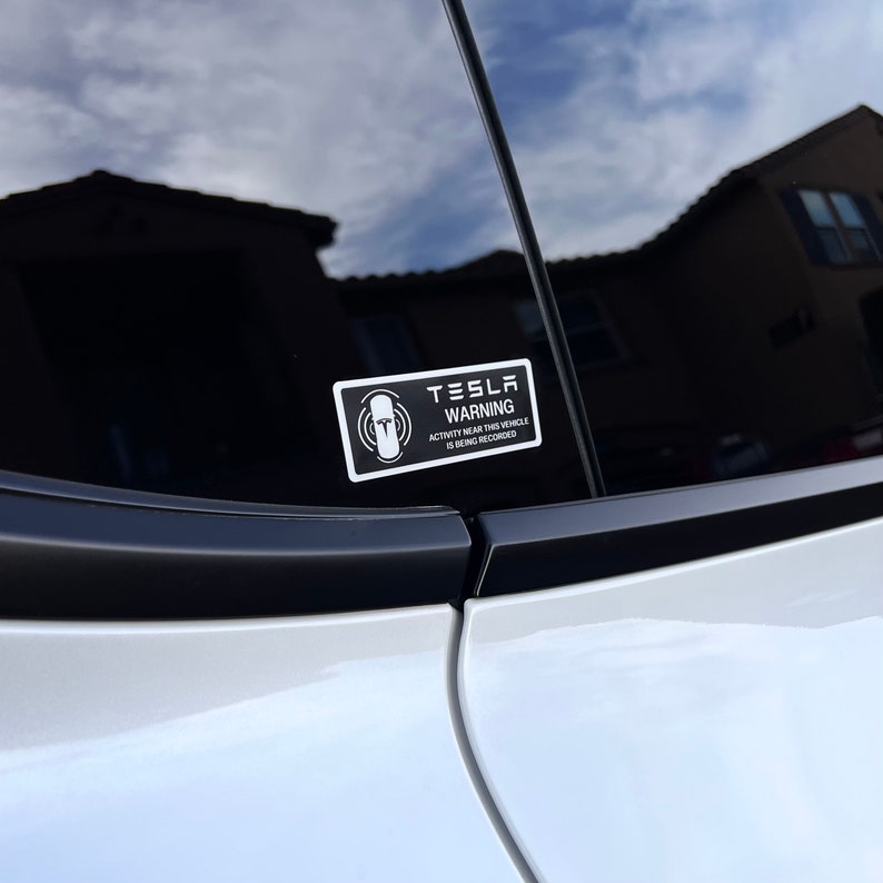 May include: A white sticker with black text on a black car window. The sticker reads "Tesla Warning Activity Near This Vehicle Is Being Recorded".